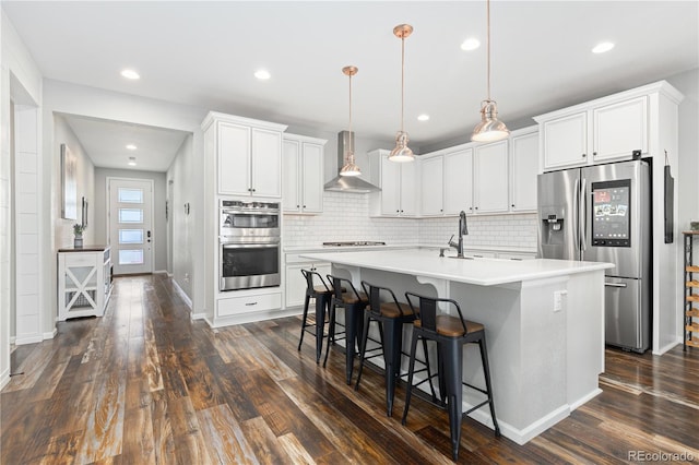 kitchen with appliances with stainless steel finishes, pendant lighting, white cabinets, a center island with sink, and wall chimney exhaust hood