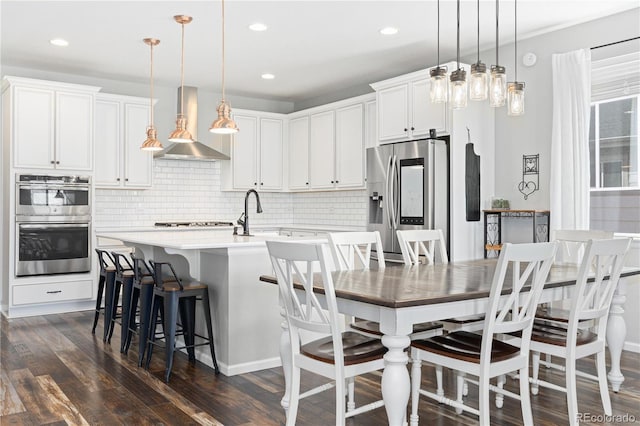 kitchen with hanging light fixtures, an island with sink, stainless steel appliances, range hood, and white cabinets