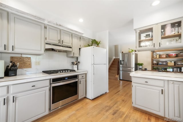 kitchen with stainless steel appliances, under cabinet range hood, light countertops, light wood-style floors, and glass insert cabinets