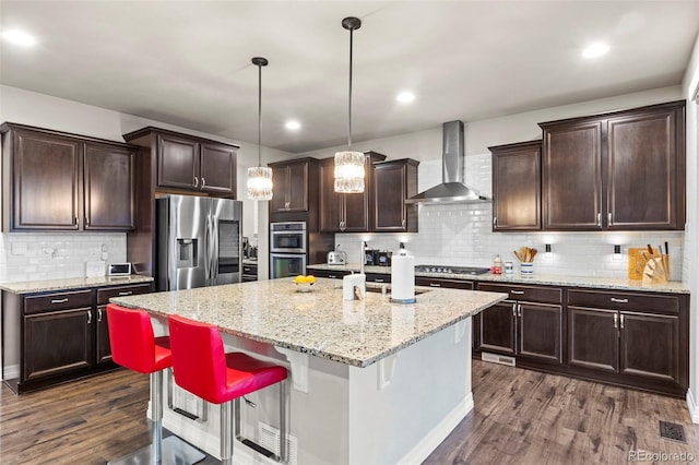 kitchen with an island with sink, dark brown cabinetry, appliances with stainless steel finishes, wall chimney exhaust hood, and dark wood-style flooring