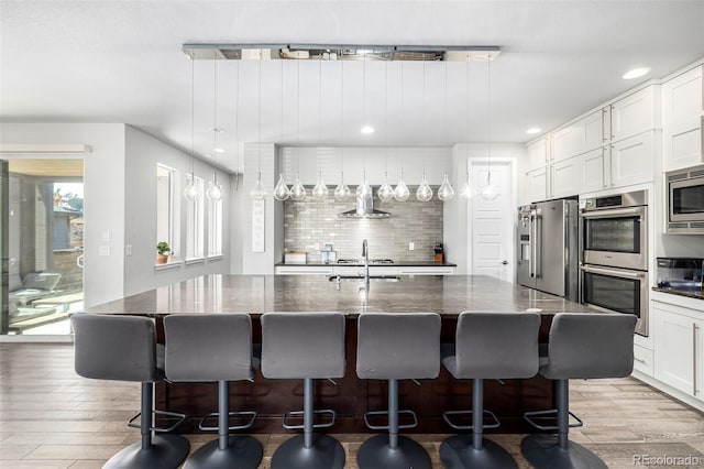 kitchen featuring stainless steel appliances, wall chimney exhaust hood, a kitchen bar, a kitchen island with sink, and pendant lighting