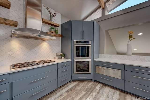 kitchen featuring wall chimney exhaust hood, vaulted ceiling with beams, light wood-type flooring, appliances with stainless steel finishes, and decorative backsplash