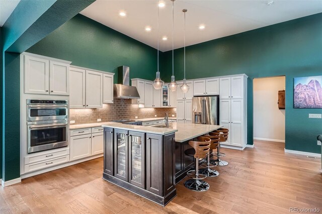 kitchen with pendant lighting, stainless steel appliances, glass insert cabinets, a kitchen island with sink, and wall chimney range hood