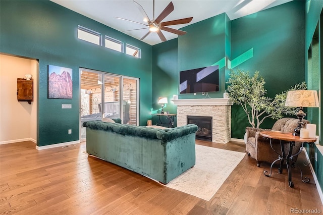 living room with a high ceiling, light wood-style floors, ceiling fan, a stone fireplace, and baseboards