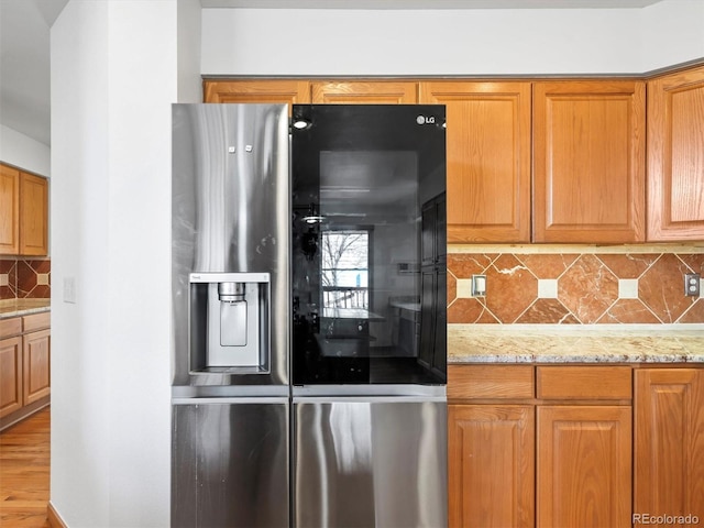 kitchen featuring decorative backsplash, light stone counters, brown cabinetry, and stainless steel refrigerator with ice dispenser