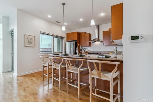 kitchen with wall chimney range hood, hanging light fixtures, stainless steel refrigerator, kitchen peninsula, and light wood-type flooring