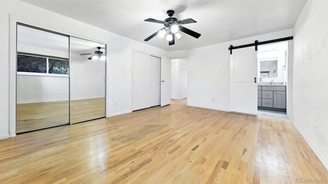 unfurnished bedroom with two closets, ceiling fan, a barn door, light hardwood / wood-style flooring, and ensuite bath