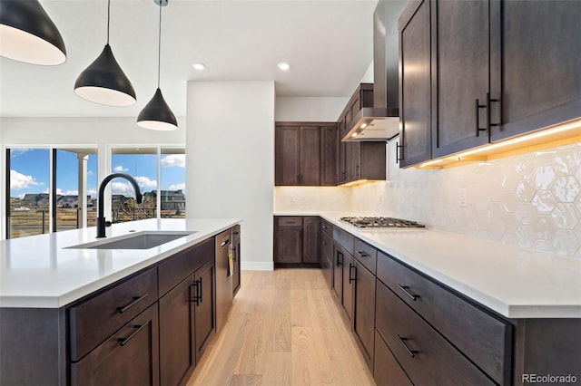 kitchen with light hardwood / wood-style floors, an island with sink, hanging light fixtures, wall chimney range hood, and sink