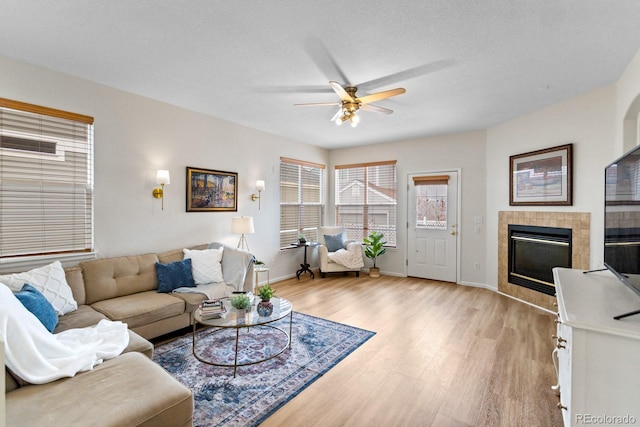 living room with ceiling fan, light wood-type flooring, a tiled fireplace, and baseboards