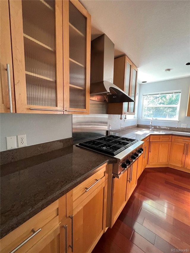 kitchen featuring wall chimney exhaust hood, dark wood finished floors, glass insert cabinets, stainless steel gas cooktop, and a sink