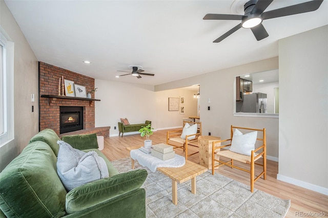 living room featuring light wood finished floors, a brick fireplace, baseboards, and ceiling fan