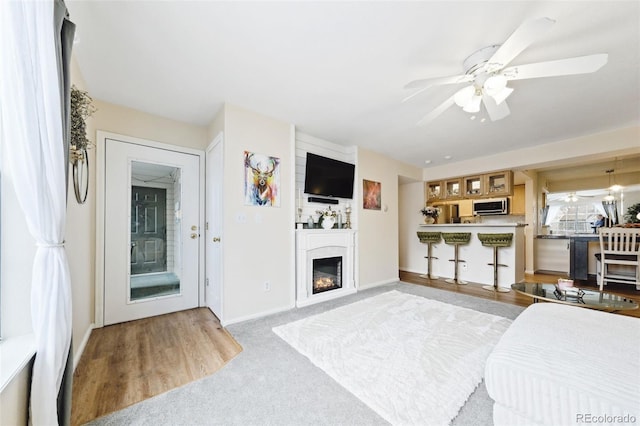 living area featuring baseboards, a glass covered fireplace, a ceiling fan, and light colored carpet