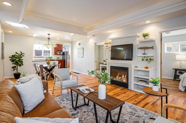 living room with a raised ceiling, a tiled fireplace, crown molding, and light wood-type flooring