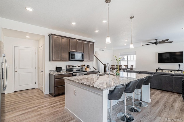 kitchen featuring sink, appliances with stainless steel finishes, a kitchen island with sink, dark brown cabinets, and light stone counters