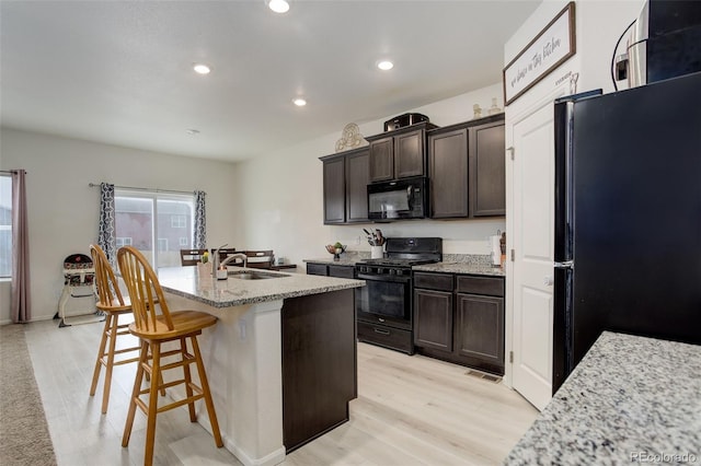 kitchen with dark brown cabinetry, a center island with sink, light stone counters, black appliances, and a sink