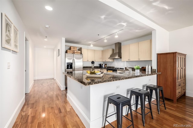 kitchen featuring light hardwood / wood-style floors, a breakfast bar area, light brown cabinets, dark stone countertops, and wall chimney range hood