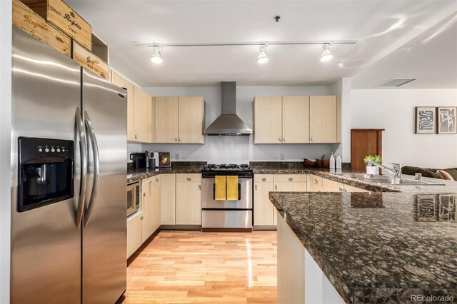 kitchen with wall chimney exhaust hood, light brown cabinets, stainless steel appliances, dark stone countertops, and sink