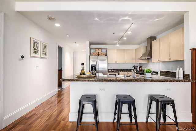 kitchen featuring stainless steel refrigerator with ice dispenser, kitchen peninsula, wall chimney exhaust hood, and dark stone countertops