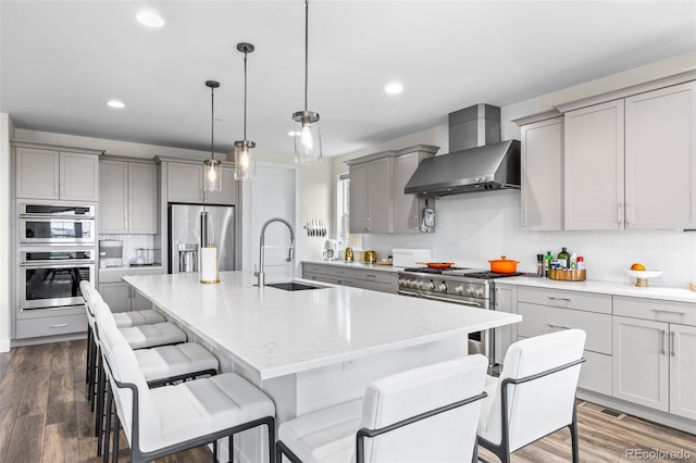 kitchen featuring a kitchen island with sink, stainless steel appliances, sink, dark wood-type flooring, and wall chimney exhaust hood