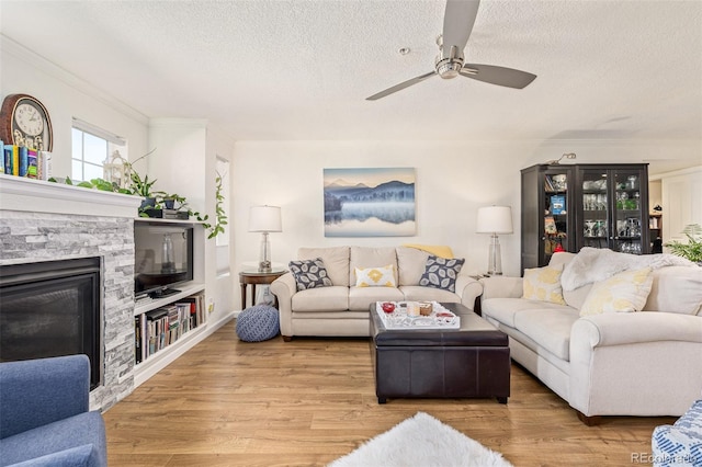 living room featuring a stone fireplace, a textured ceiling, light hardwood / wood-style floors, ceiling fan, and built in shelves