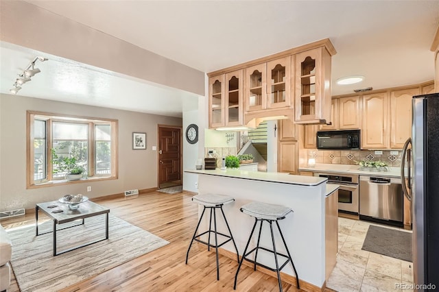 kitchen featuring a breakfast bar area, light brown cabinetry, glass insert cabinets, appliances with stainless steel finishes, and tasteful backsplash