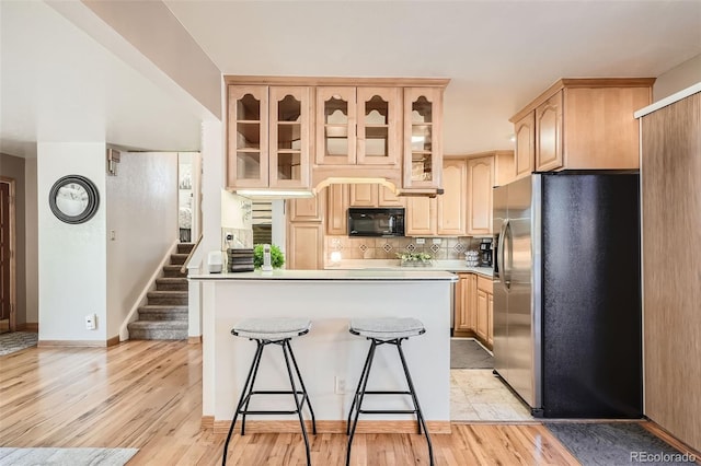 kitchen with light brown cabinetry, stainless steel fridge, glass insert cabinets, black microwave, and light wood-type flooring