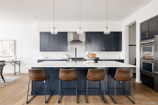 kitchen featuring light wood finished floors, light countertops, stainless steel double oven, wall chimney range hood, and a kitchen bar