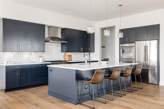 kitchen featuring light wood finished floors, wall chimney exhaust hood, a kitchen island with sink, stainless steel appliances, and a sink