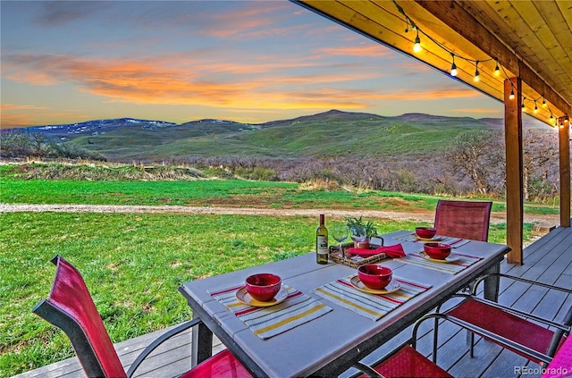 deck at dusk with a lawn, outdoor dining area, and a mountain view