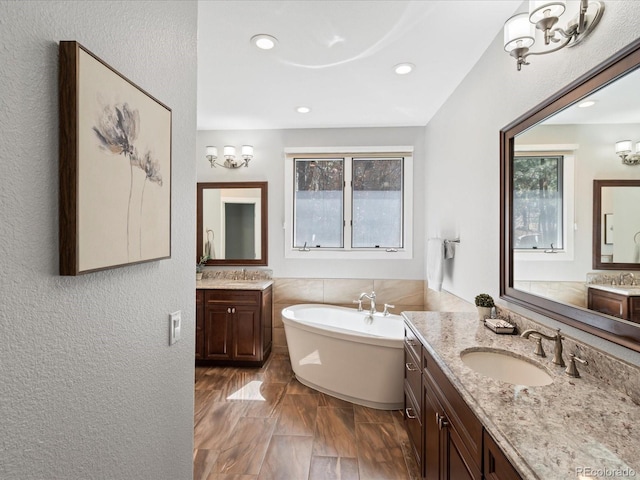 bathroom featuring a soaking tub, plenty of natural light, a textured wall, two vanities, and recessed lighting