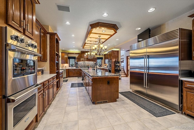 kitchen with appliances with stainless steel finishes, backsplash, a center island with sink, an inviting chandelier, and decorative light fixtures