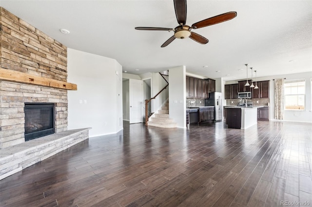 unfurnished living room featuring a ceiling fan, a fireplace, dark wood finished floors, baseboards, and stairs