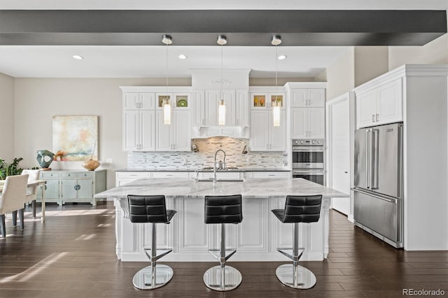 kitchen with pendant lighting, a center island with sink, appliances with stainless steel finishes, glass insert cabinets, and white cabinetry