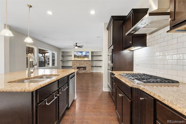 kitchen with pendant lighting, sink, stainless steel appliances, wall chimney range hood, and a stone fireplace