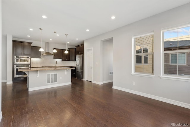 kitchen with wall chimney exhaust hood, pendant lighting, an island with sink, a breakfast bar, and appliances with stainless steel finishes