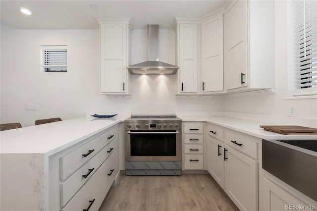 kitchen featuring light wood finished floors, white cabinets, high end stove, a peninsula, and wall chimney exhaust hood