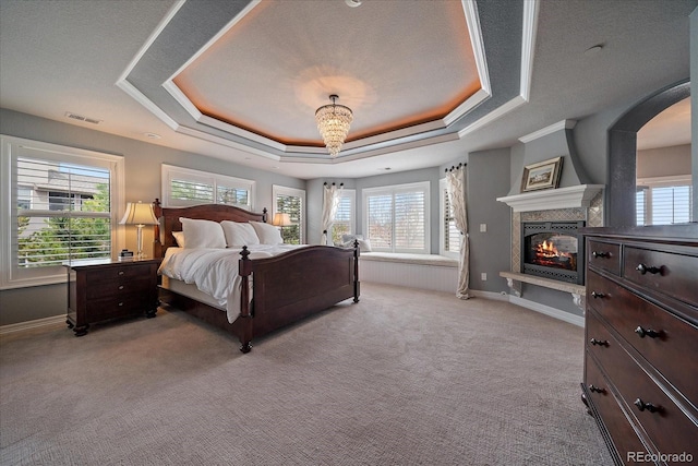 bedroom with a tray ceiling, a glass covered fireplace, visible vents, arched walkways, and light carpet
