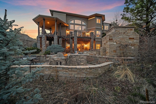 back of house at dusk featuring a balcony, area for grilling, a patio area, stucco siding, and stone siding