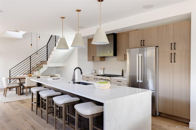 kitchen with high end refrigerator, a kitchen island with sink, sink, wall chimney range hood, and pendant lighting