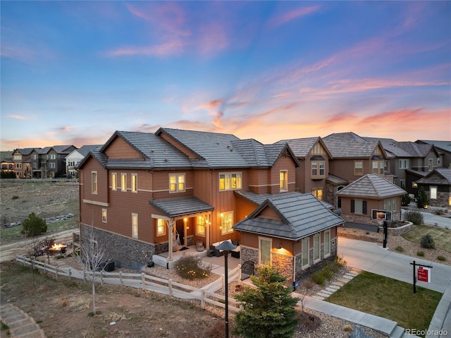 back of house at dusk featuring driveway, stone siding, a tiled roof, fence, and a residential view