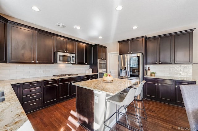 kitchen with light stone counters, dark wood-style flooring, stainless steel appliances, dark brown cabinets, and a kitchen bar