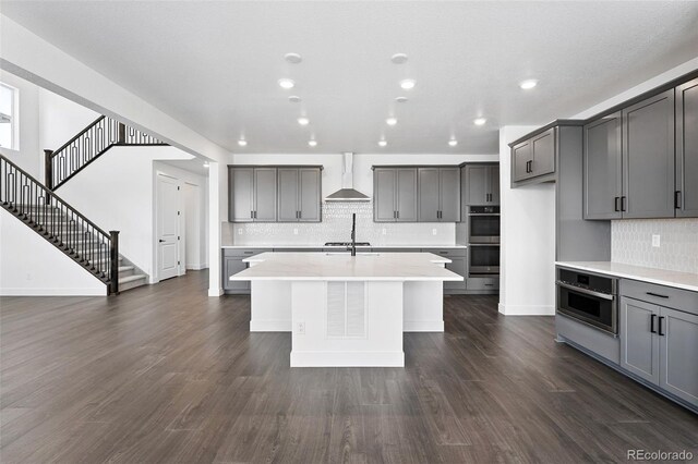 16565 Black Rose Cir, Parker, CO 80134 - kitchen featuring wall chimney exhaust hood, double oven, gray cabinets, a kitchen island with sink, and decorative backsplash
