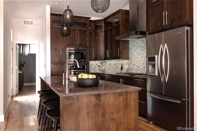 kitchen featuring stainless steel appliances, wall chimney range hood, light wood-style floors, and tasteful backsplash