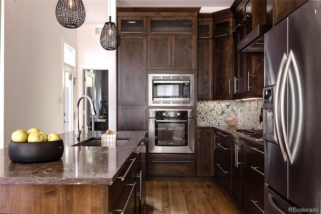 kitchen featuring appliances with stainless steel finishes, a sink, wood finished floors, dark stone counters, and glass insert cabinets