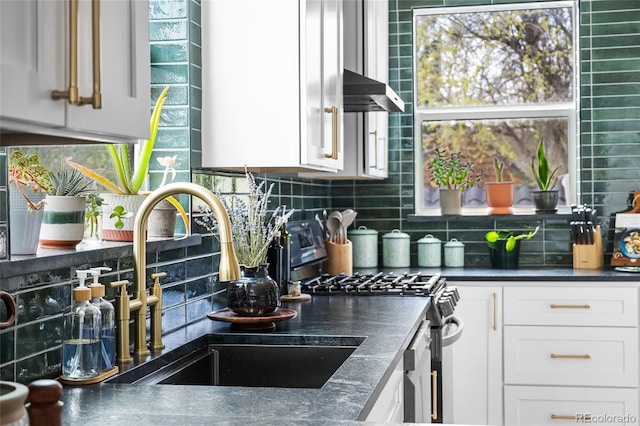 kitchen with wall chimney range hood, backsplash, dark countertops, stainless steel gas stove, and a sink