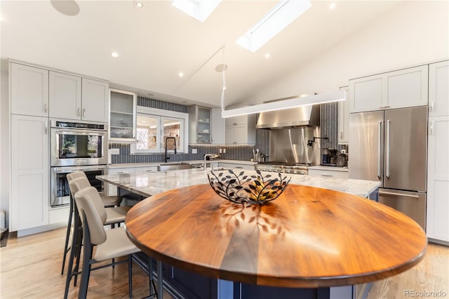 kitchen with a sink, lofted ceiling with skylight, backsplash, wall chimney range hood, and appliances with stainless steel finishes