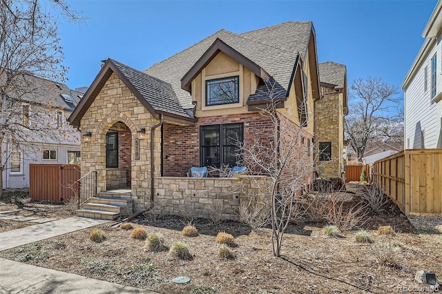 tudor house featuring brick siding, stone siding, a shingled roof, and fence