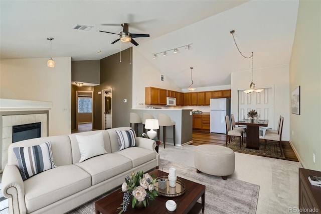 living room featuring baseboards, visible vents, a ceiling fan, a tiled fireplace, and high vaulted ceiling