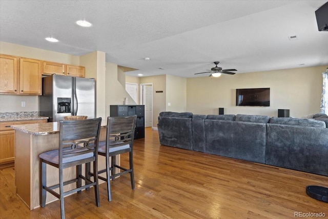 kitchen with light stone counters, stainless steel fridge with ice dispenser, light brown cabinets, wood finished floors, and a kitchen bar