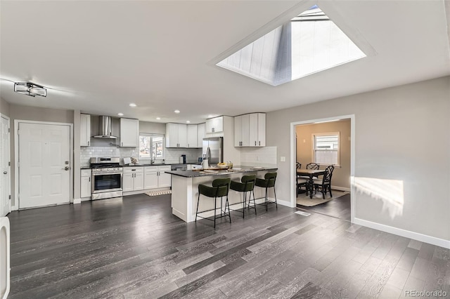 kitchen with dark countertops, decorative backsplash, appliances with stainless steel finishes, a peninsula, and wall chimney exhaust hood